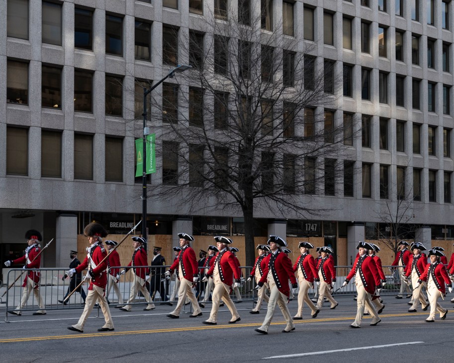 Military band members in red uniforms and tricorn hats marching in formation during Biden's inauguration ceremony