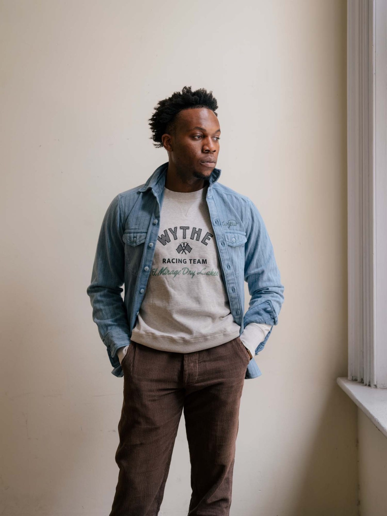 Man wearing Wythe denim shirt, graphic t-shirt and brown corduroy trousers in neutral studio setting