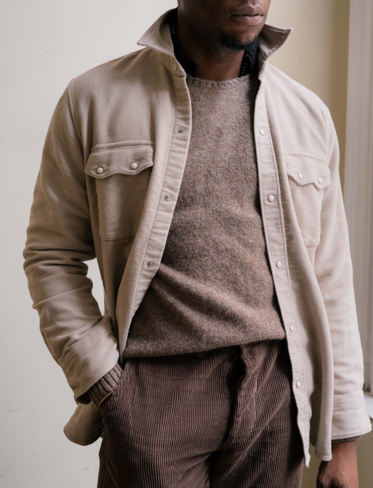 Model wearing Wythe's pearl-snap western shirt in tan cotton with brown knitwear and ribbed trousers.