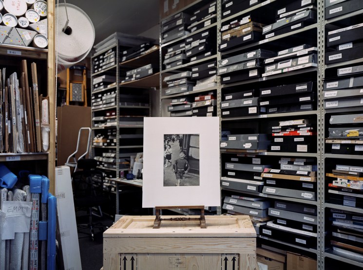 Black and white photograph displayed on easel in archival storage room with labeled film canisters on shelves