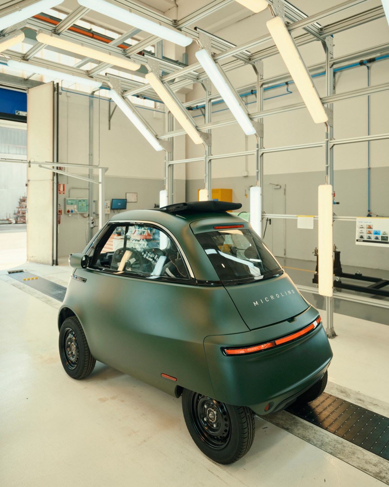 A matte-green Microlino electric car positioned on a testing platform inside a bright industrial facility.