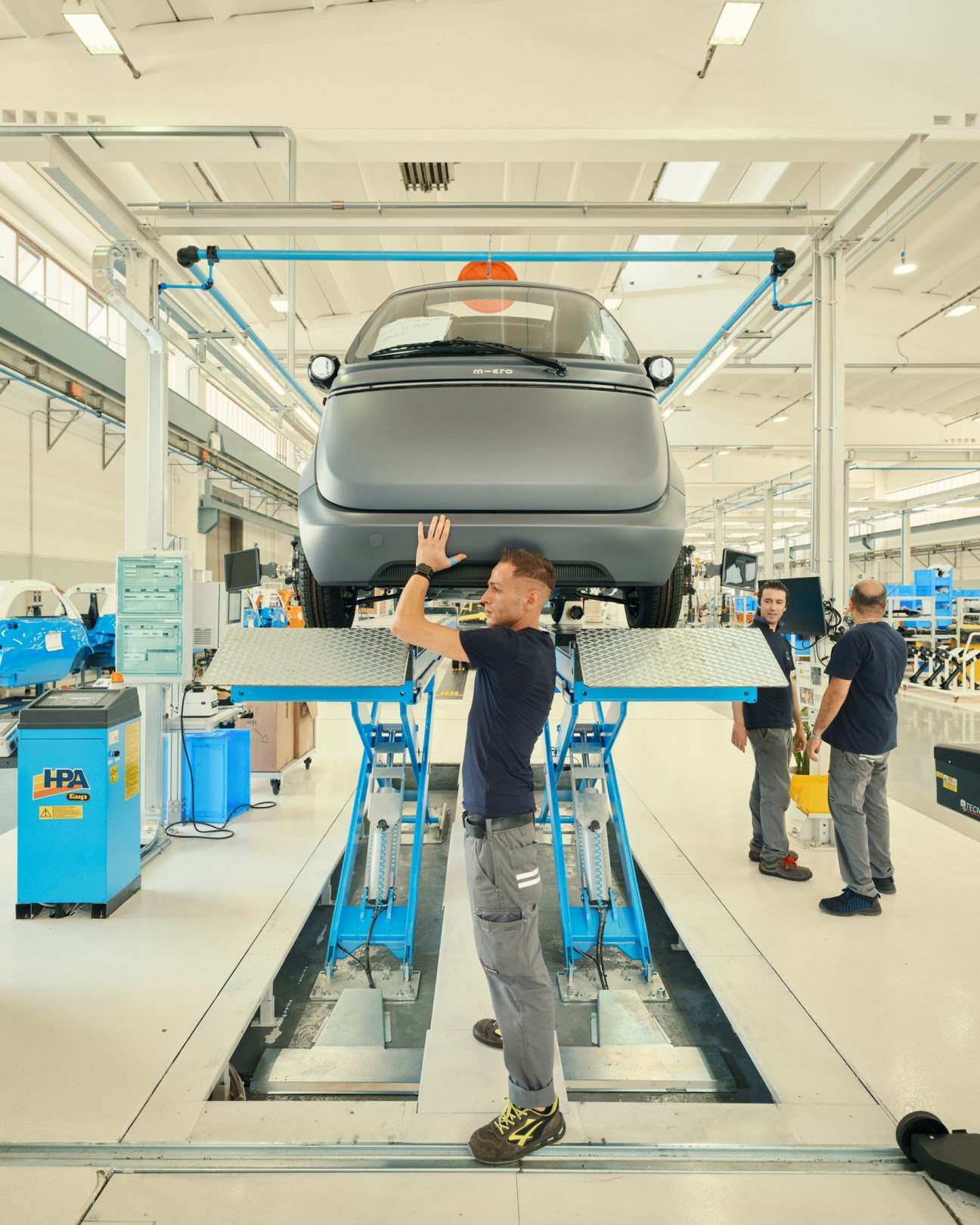 Worker inspects underside of elevated Microlino electric car on blue hydraulic lift in Turin factory