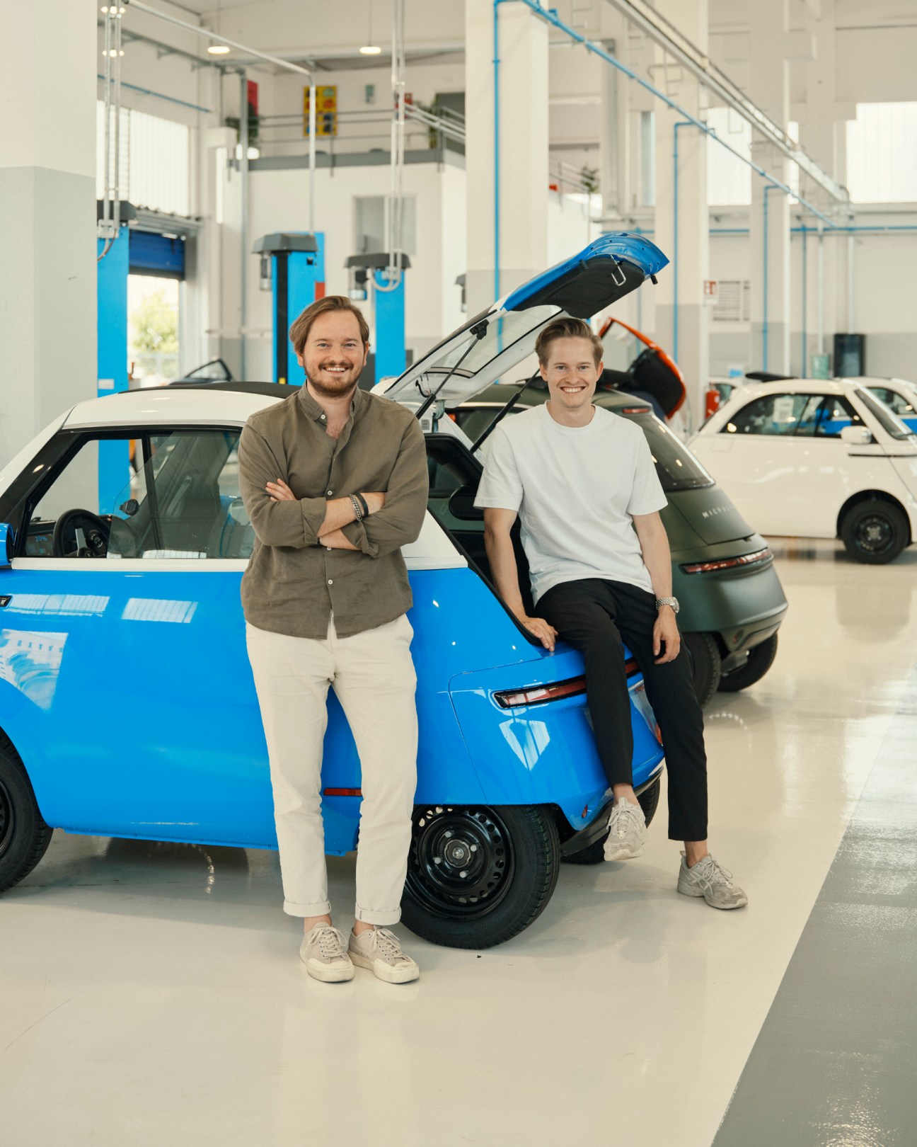 Two men standing beside a bright blue Microlino electric vehicle in a modern automotive factory