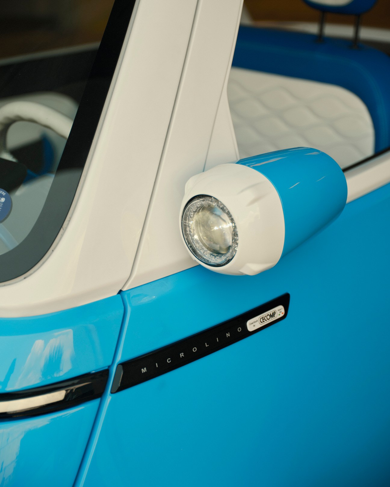Close-up of a bright blue and white Microlino electric car showing the distinctive round wing mirror and headlight