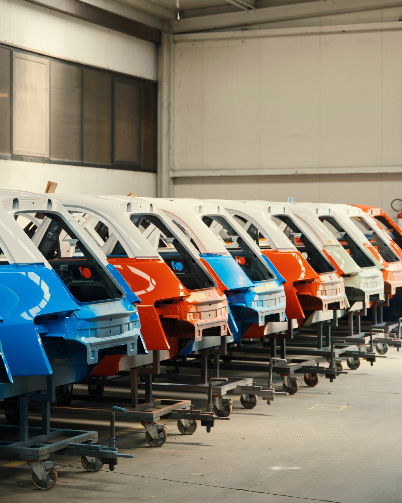 Row of colorful Microlino car bodies in blue, orange and white lined up on wheeled stands in Turin factory