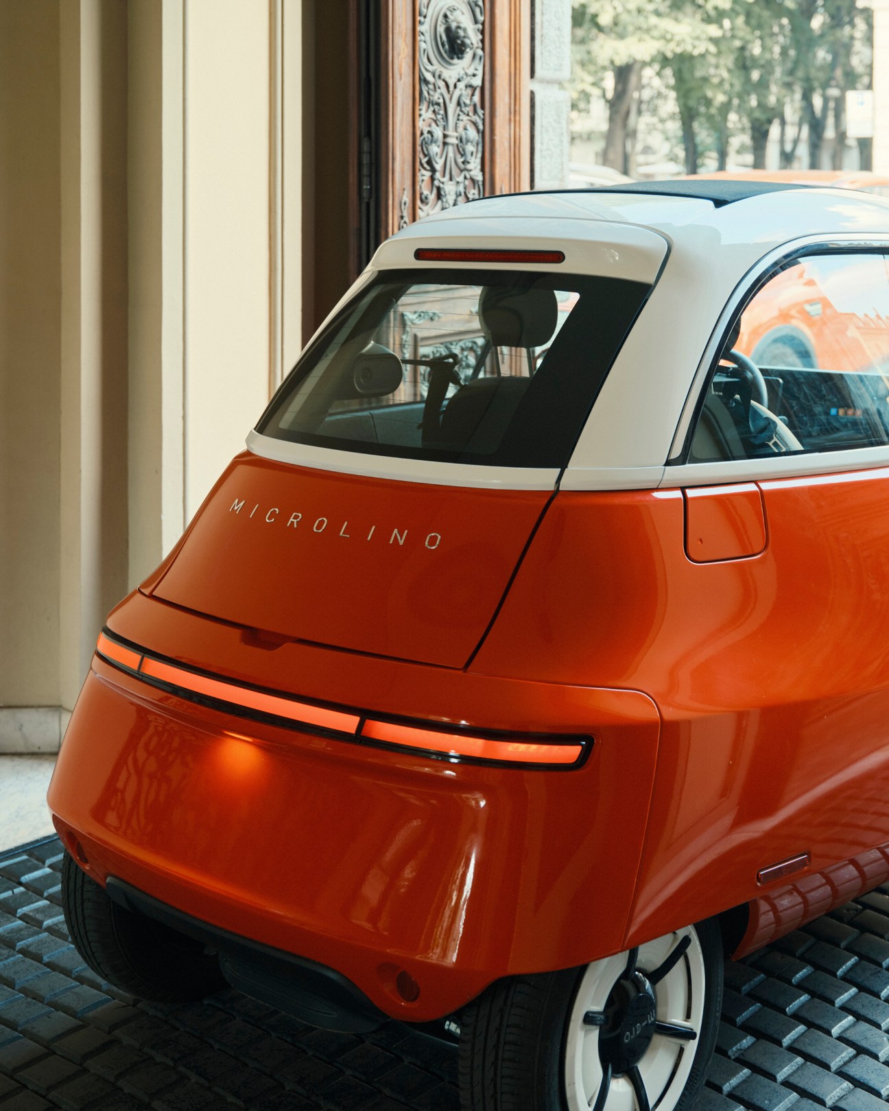Orange and white Microlino electric vehicle parked on cobblestones outside an ornate building entrance