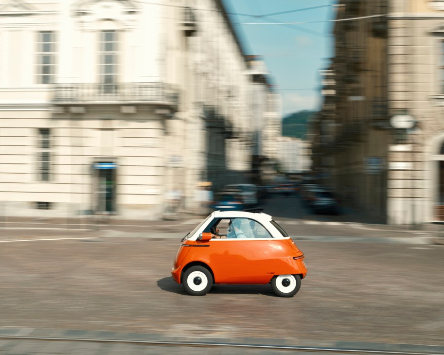 Orange Microlino electric microcar driving through European city streets with motion blur effect