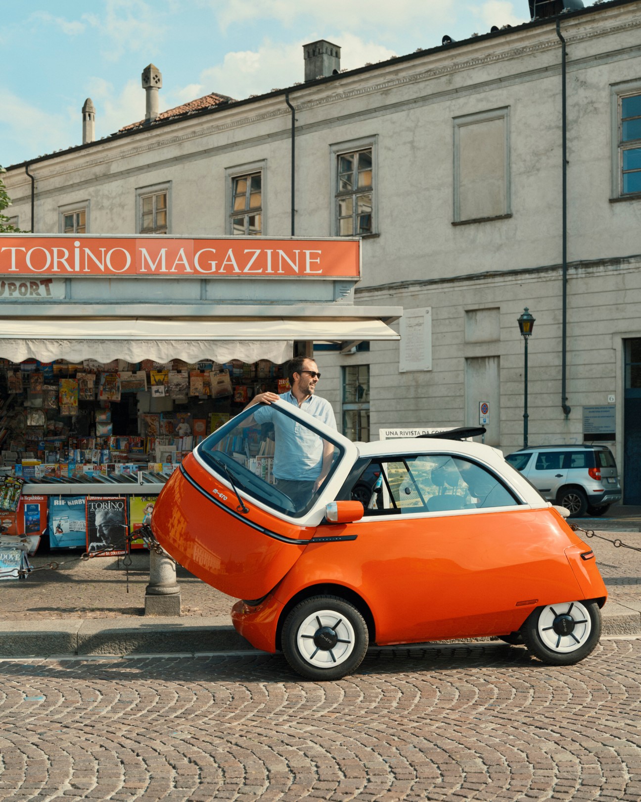 Orange Microlino electric car parked on cobblestone street in front of magazine kiosk in Turin, Italy