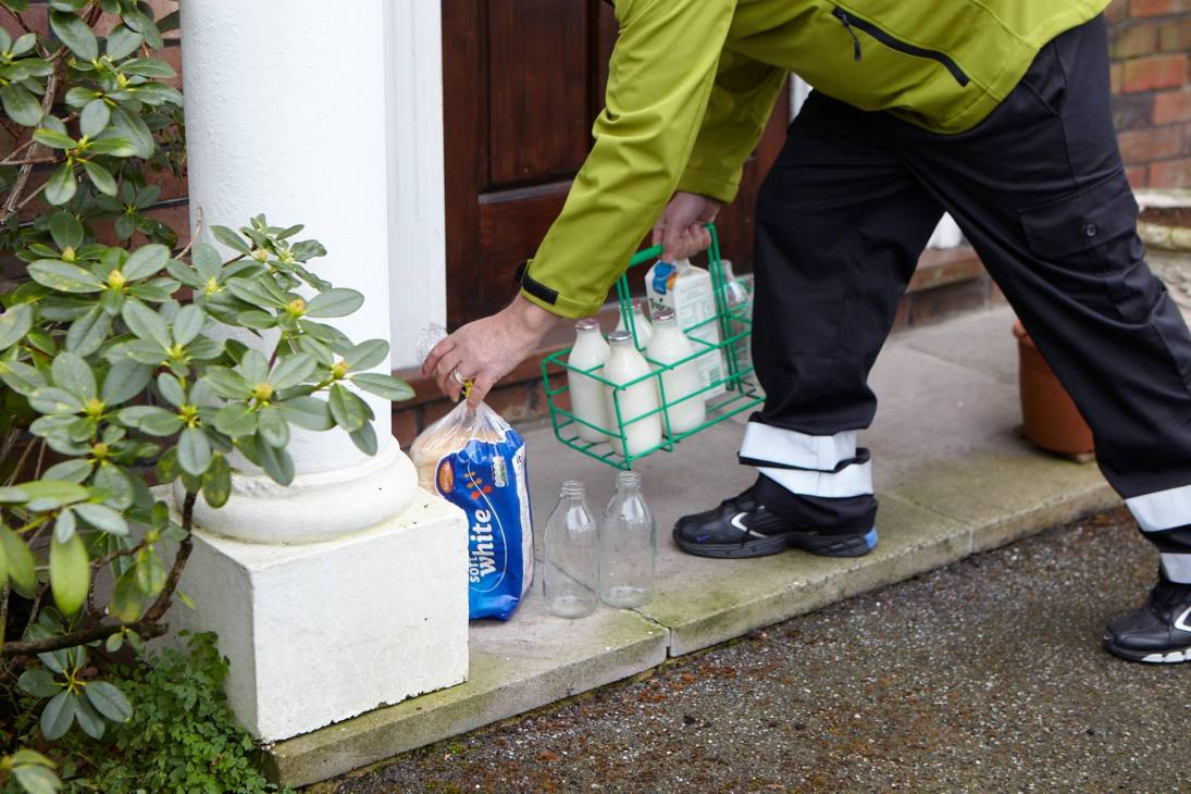 Milk delivery person in high-vis jacket placing glass milk bottles on doorstep from wire crate