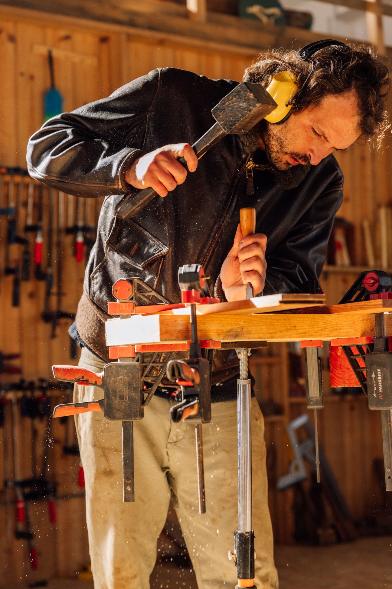 Man in black jacket using chisel and hammer on wood piece secured in workshop clamps