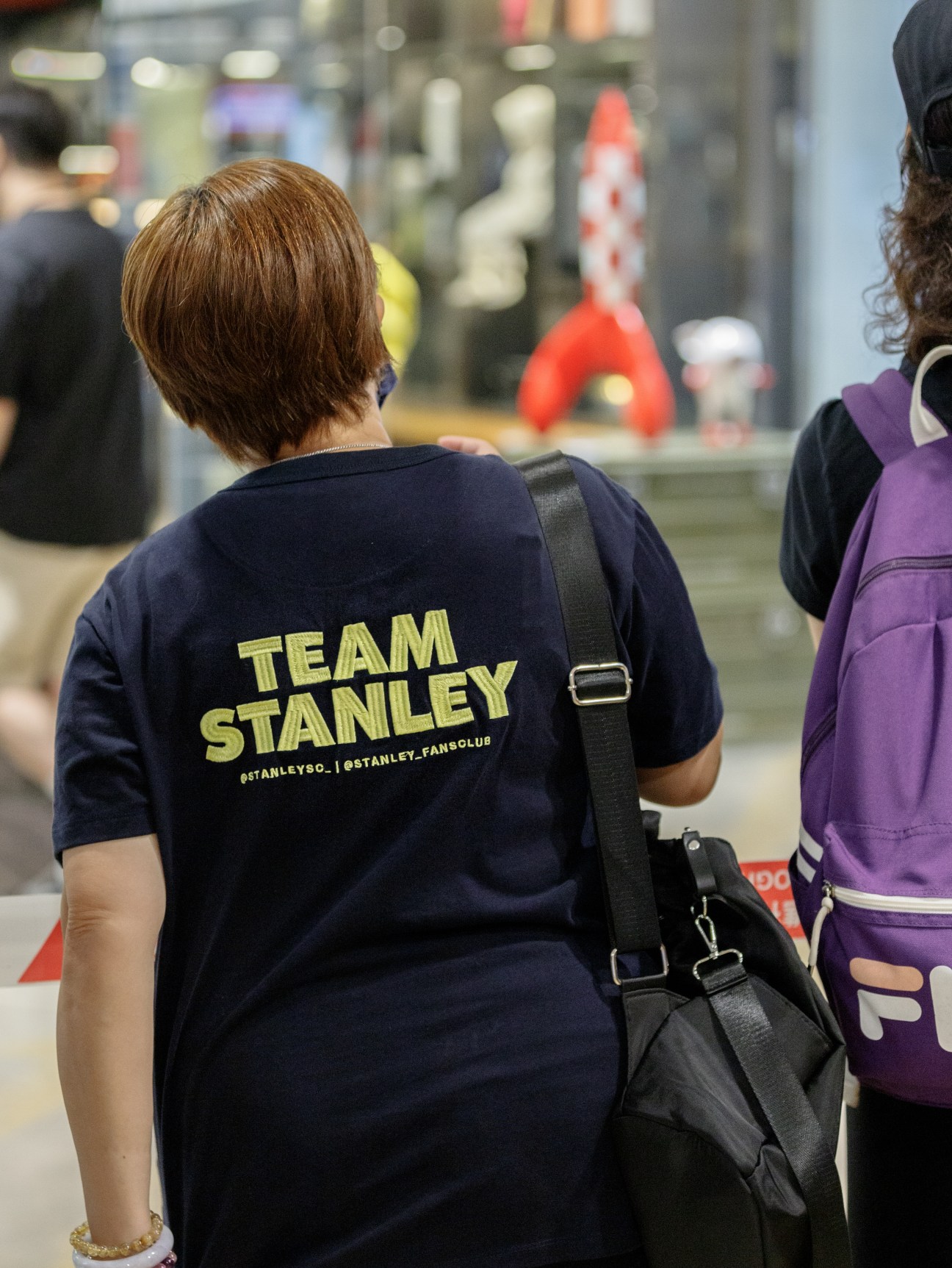 Person wearing Team Stanley t-shirt with black bag strap walking in what appears to be a shopping mall