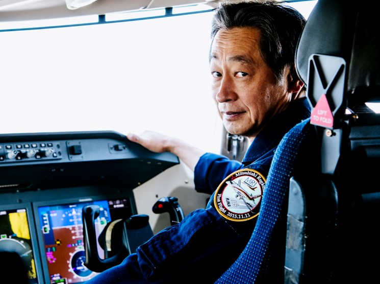 A pilot in a blue uniform with mission patches looks back from an aircraft cockpit surrounded by flight instruments.