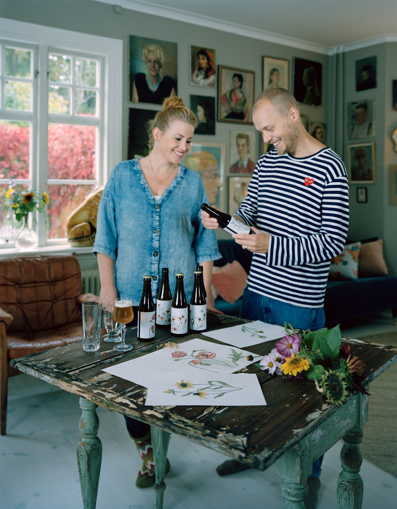 Patrik and Karolina Svensk examining a bottle of Munklägret craft beer in their home surrounded by portraits and botanical drawings.