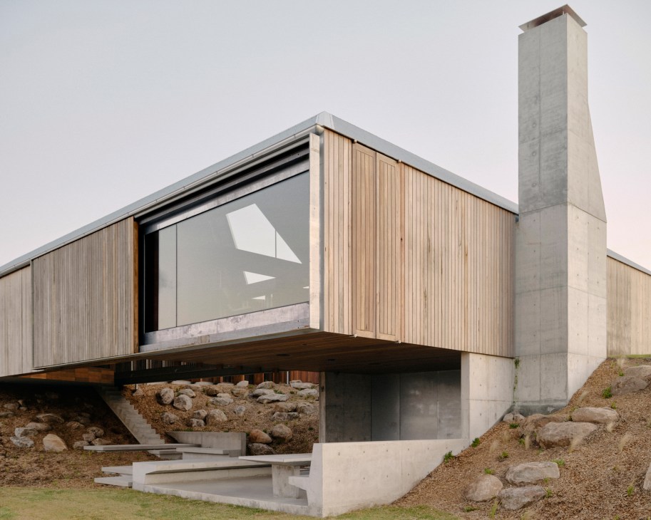 Modern house with timber cladding and concrete elements cantilevering over a sand dune with rocky landscape