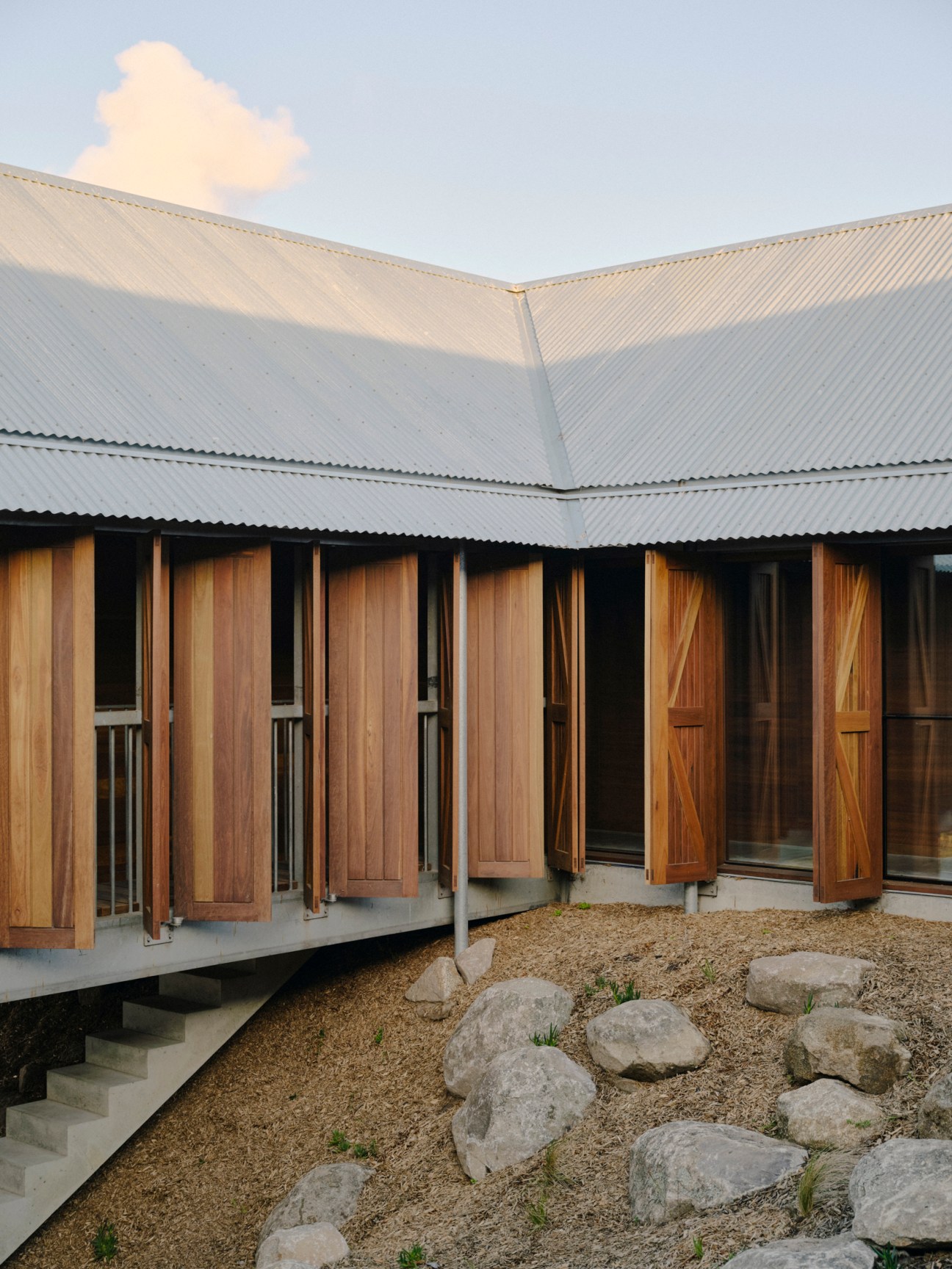 Courtyard view of farmhouse showing timber shutters, concrete structure, and landscaped ground with scattered rocks