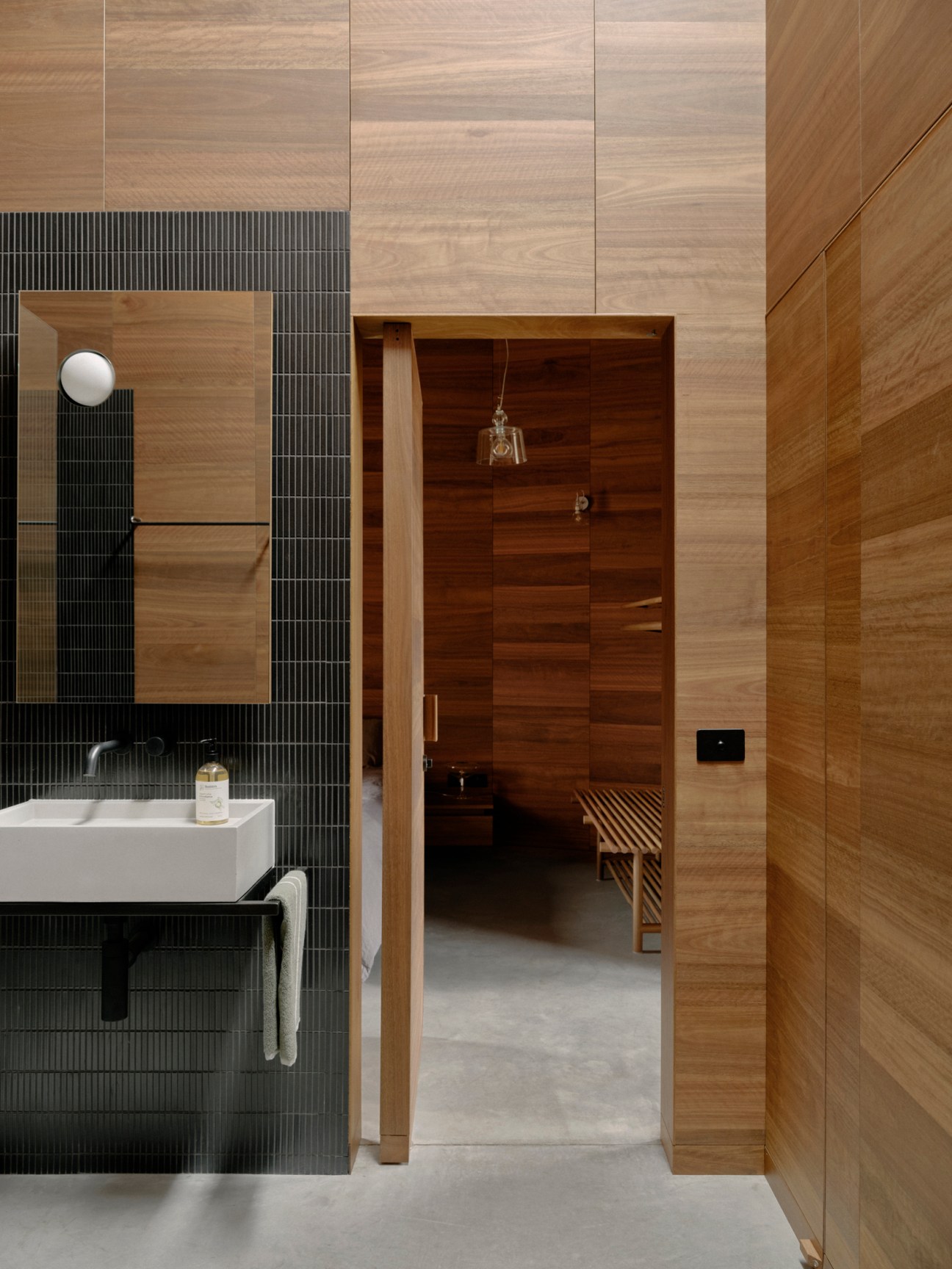 Modern bathroom interior with light wood paneling, dark mosaic tiles, white sink, and sauna entrance