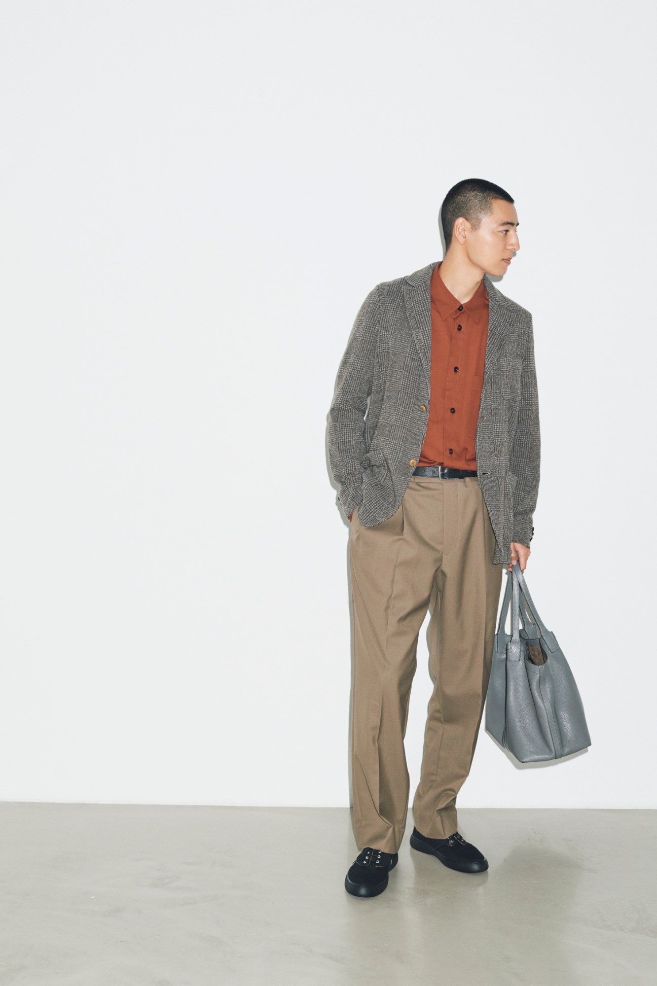 Model in grey blazer, rust shirt, and beige trousers holding leather bag against white background