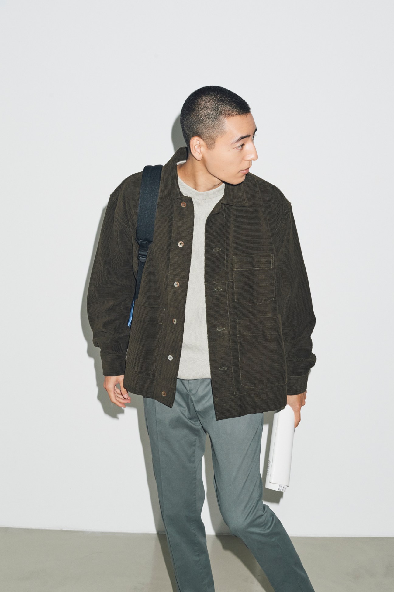 Model in profile wearing olive corduroy jacket over cream sweater with grey trousers and navy backpack against white wall