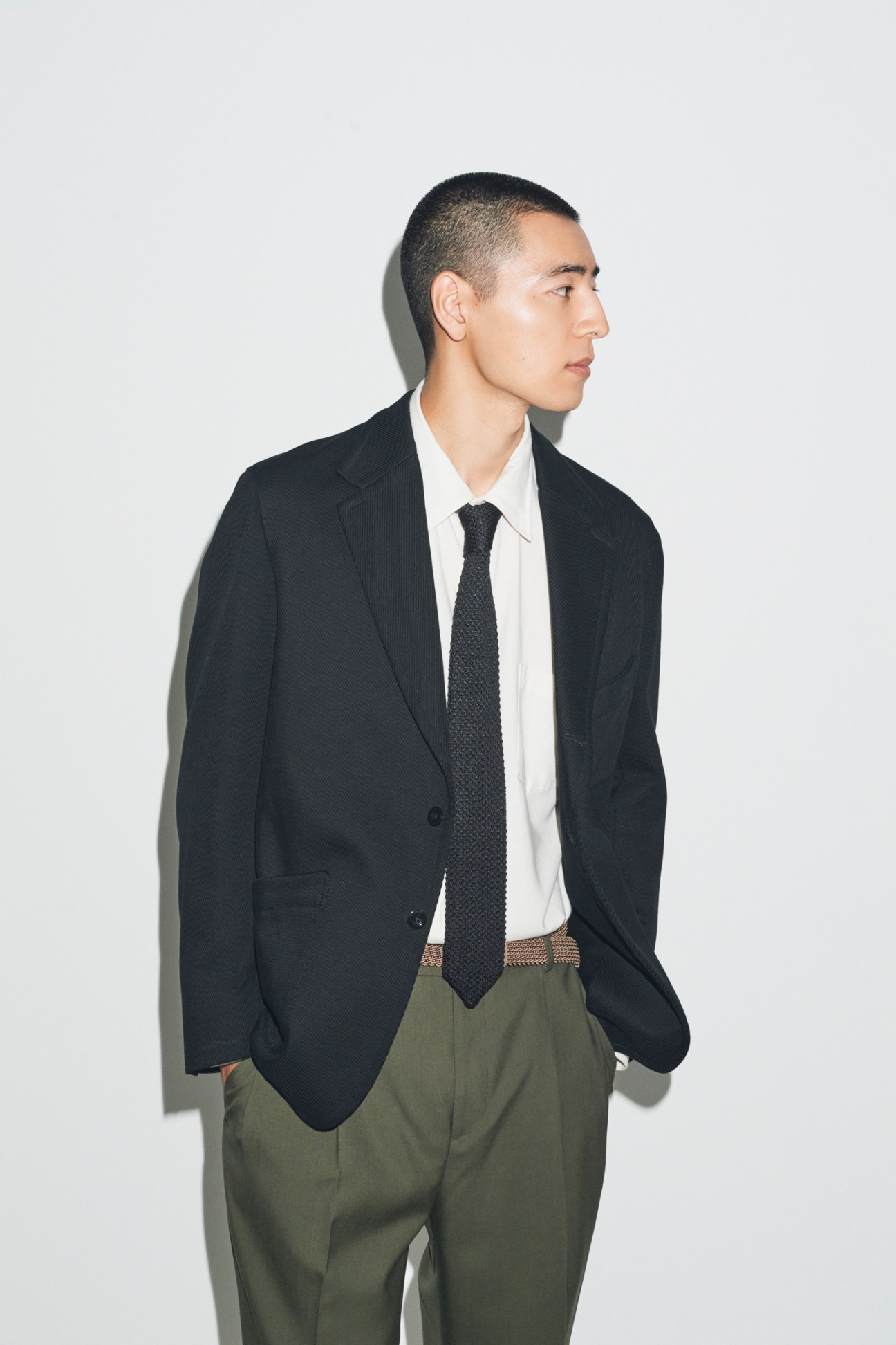 Model in profile wearing black blazer, white shirt, dark tie and olive green trousers against white background