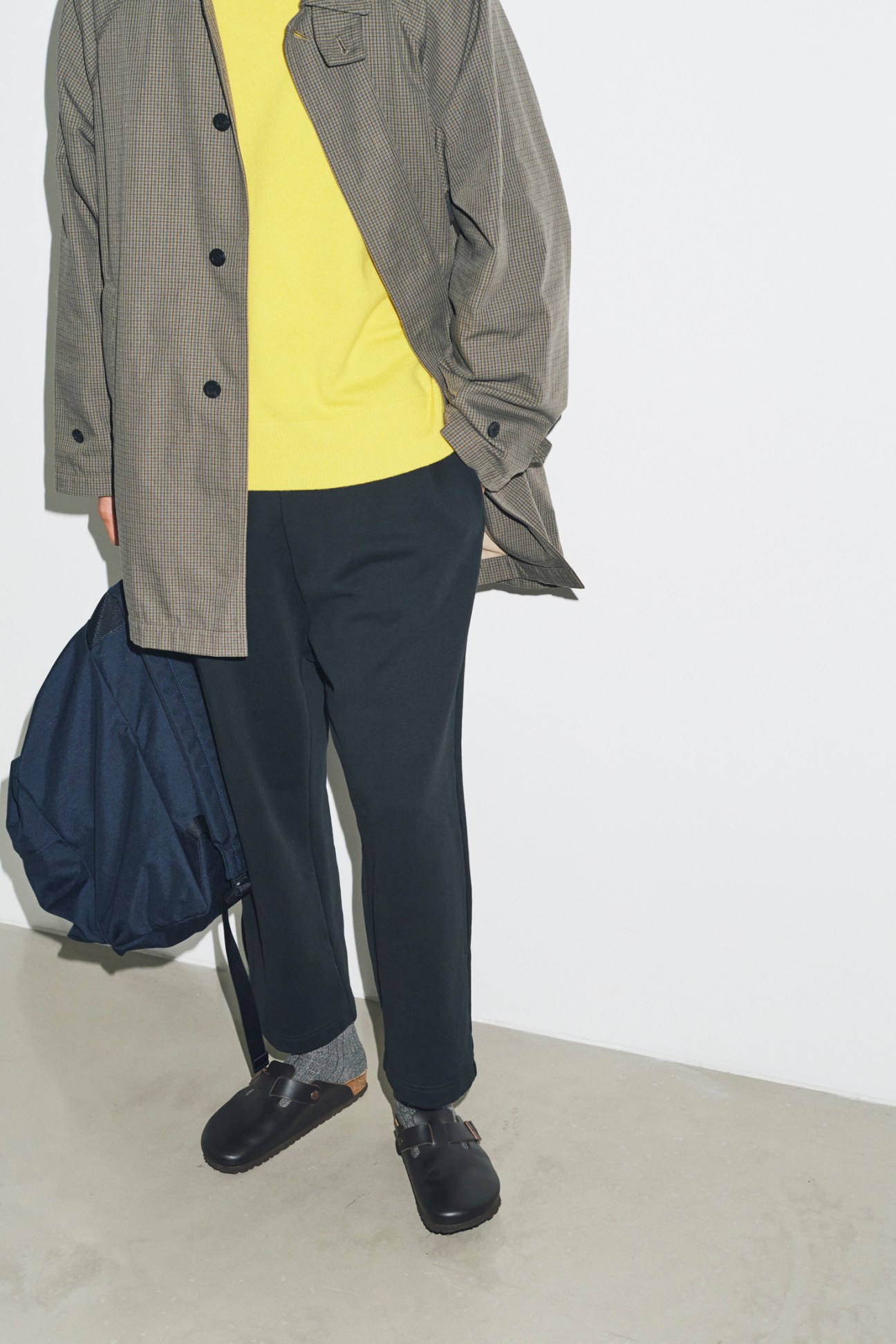 Model wearing grey checked coat over yellow jumper with navy trousers and Birkenstock sandals, carrying blue backpack