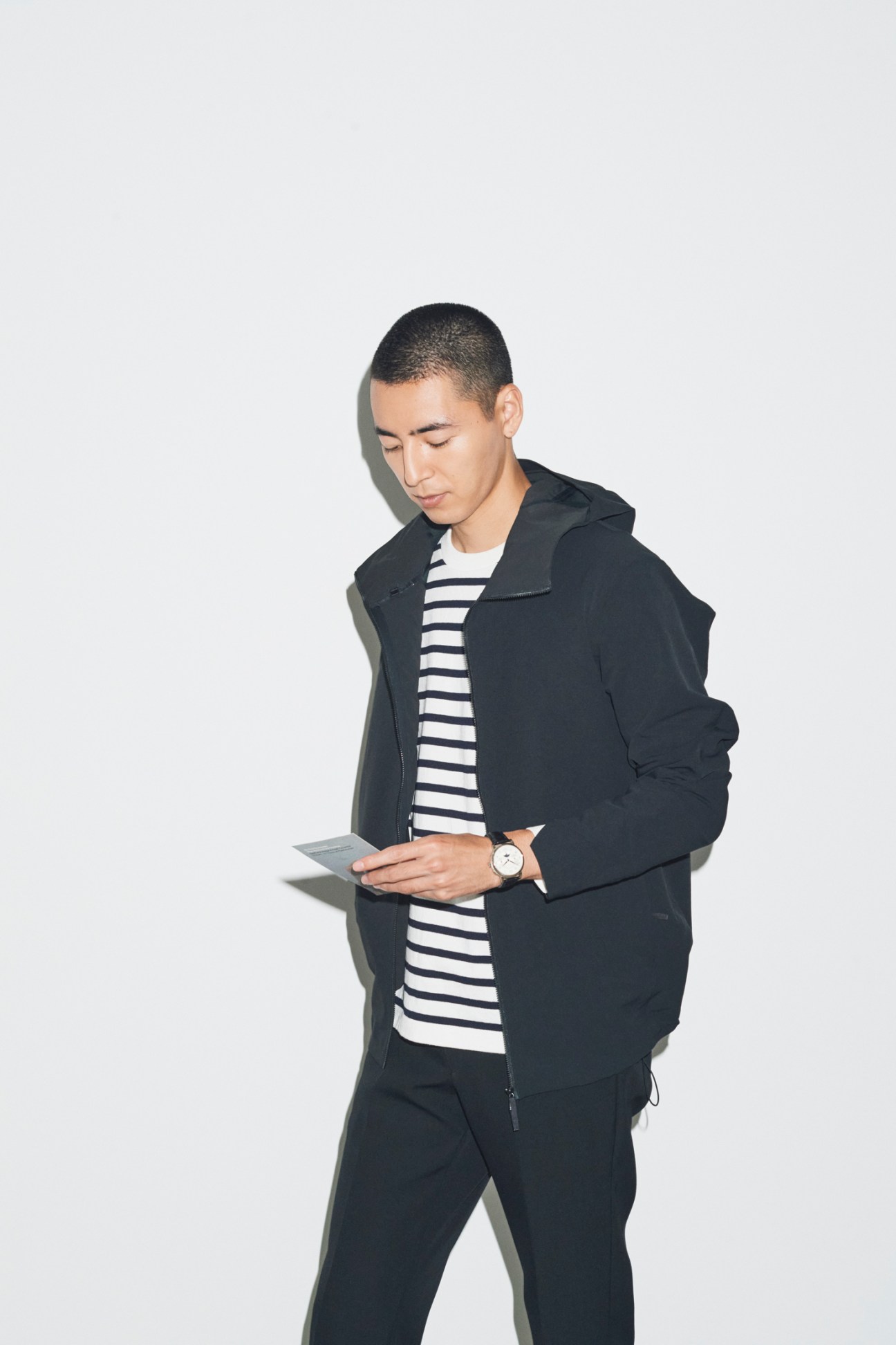 Man in navy jacket over striped shirt looking down at phone, wearing watch against white background