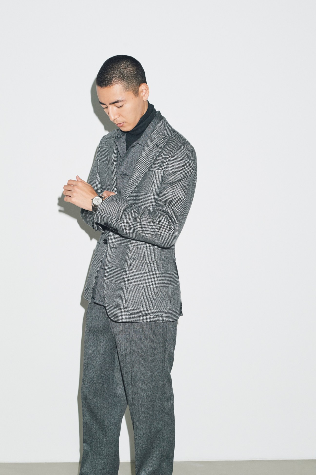 Model wearing grey glen plaid blazer over black turtleneck with grey trousers, looking down pensively and displaying watch on his wrist