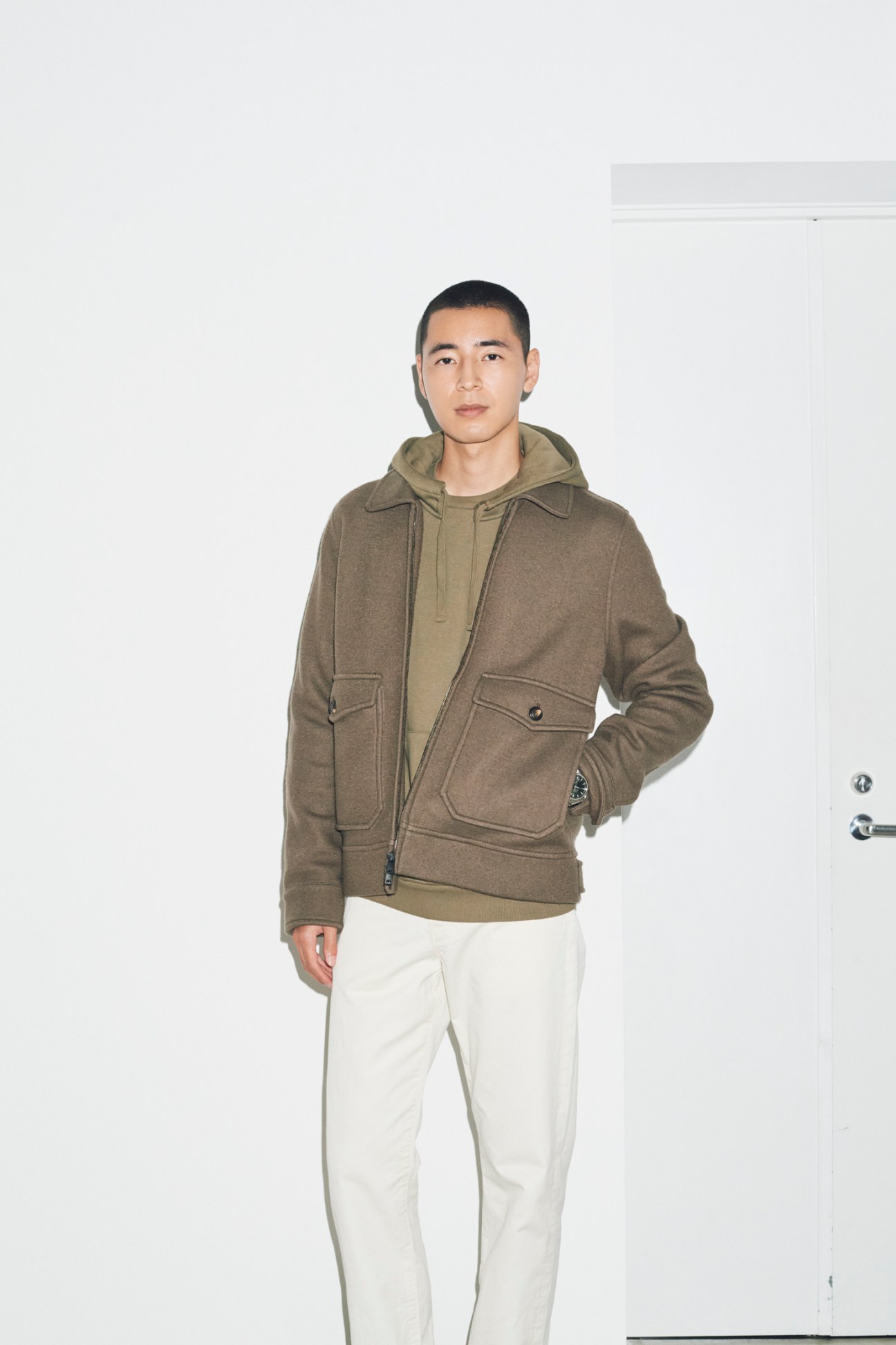 Man in olive green zip-up jacket over hoodie and white trousers against white background
