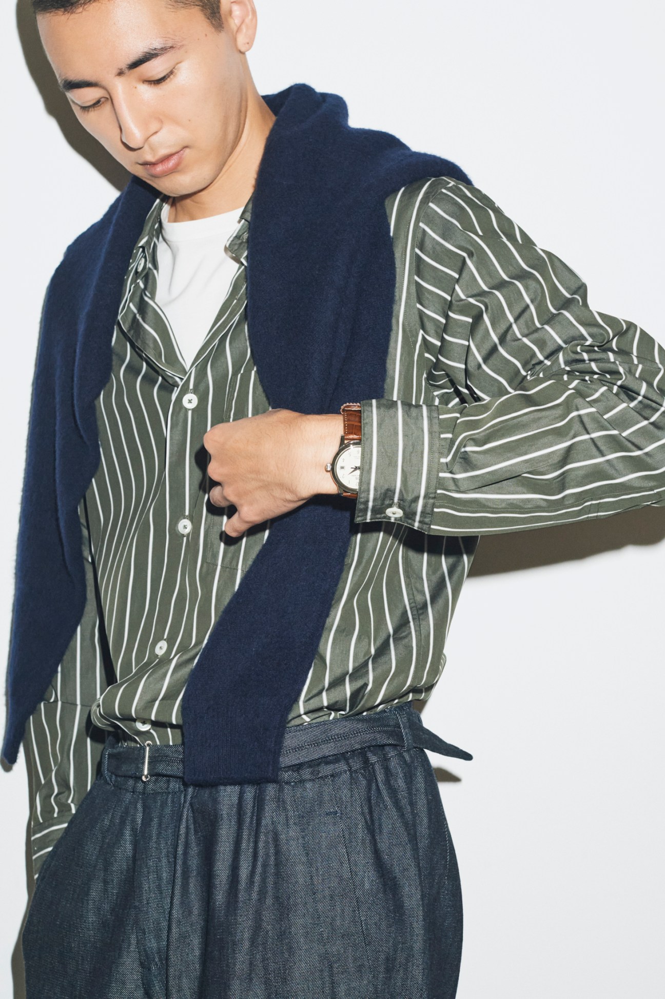 Model wearing navy jumper over striped olive shirt with brown leather watch, demonstrating autumn layering techniques