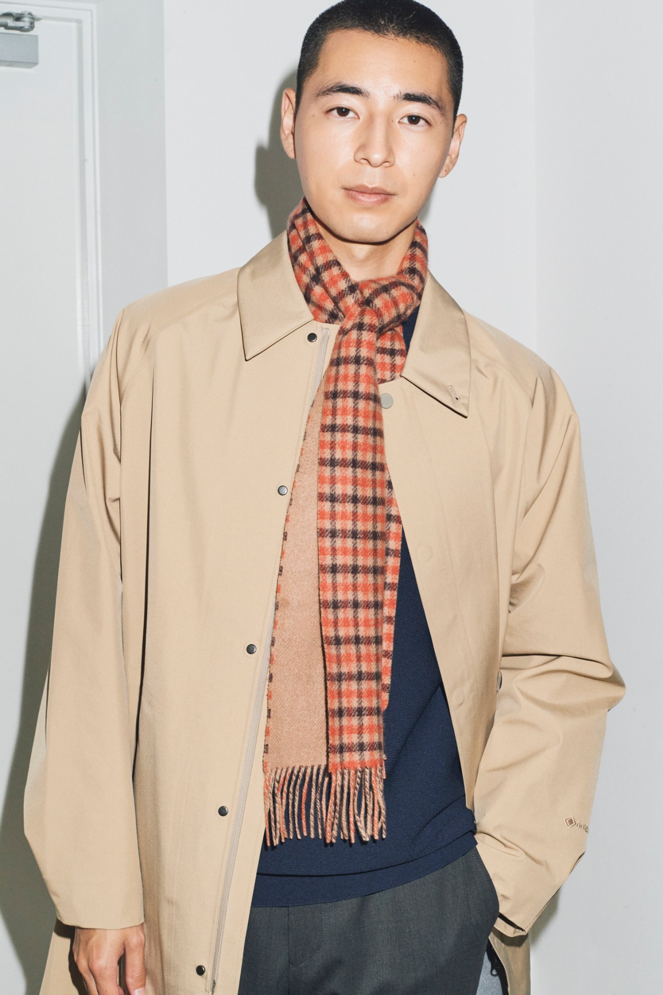 Man in beige coat with checkered scarf and navy sweater demonstrating autumn layering against white wall