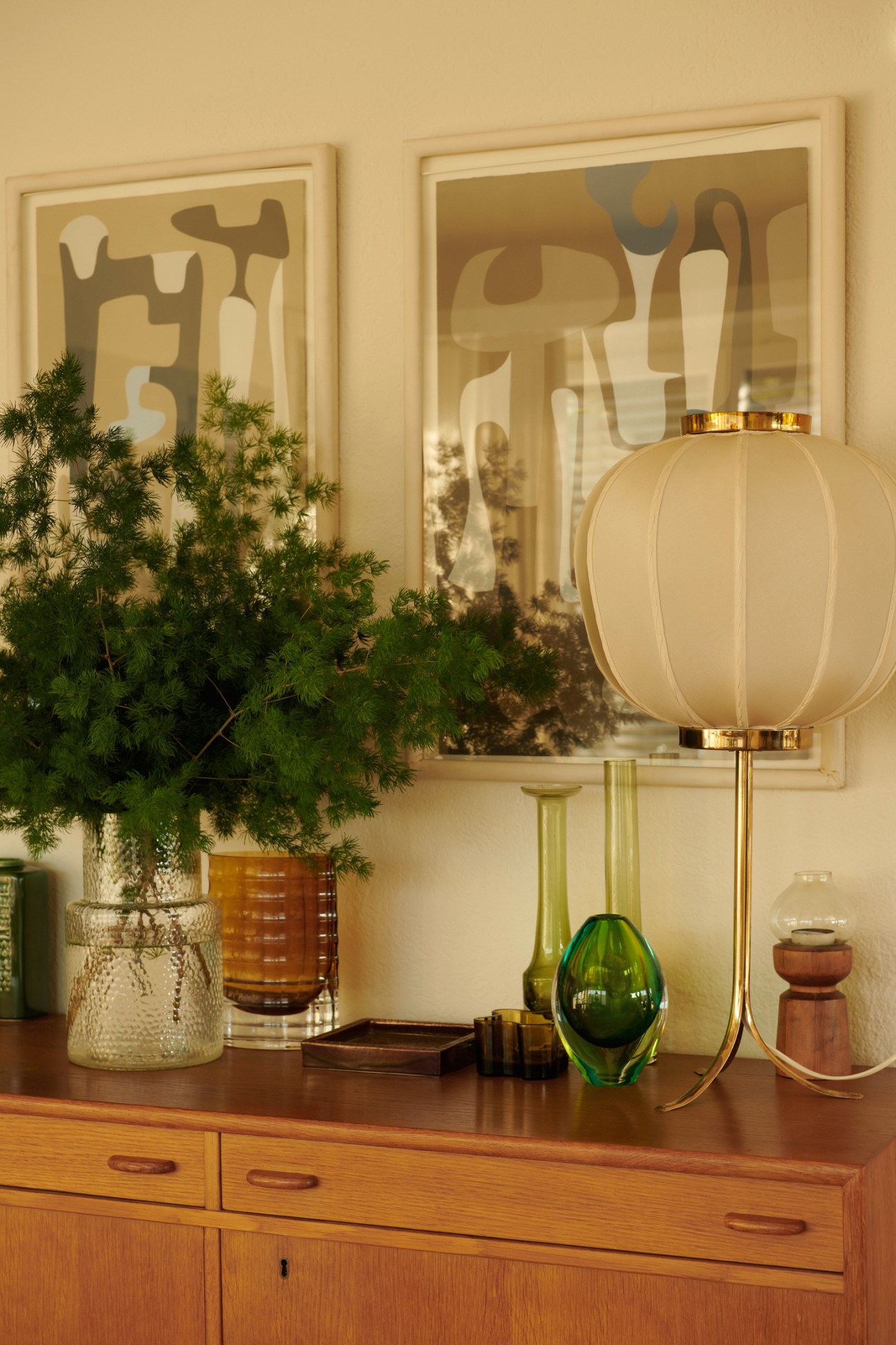 Modernist abstract artworks in frames displayed on wall above wooden sideboard with table lamp and decorative objects