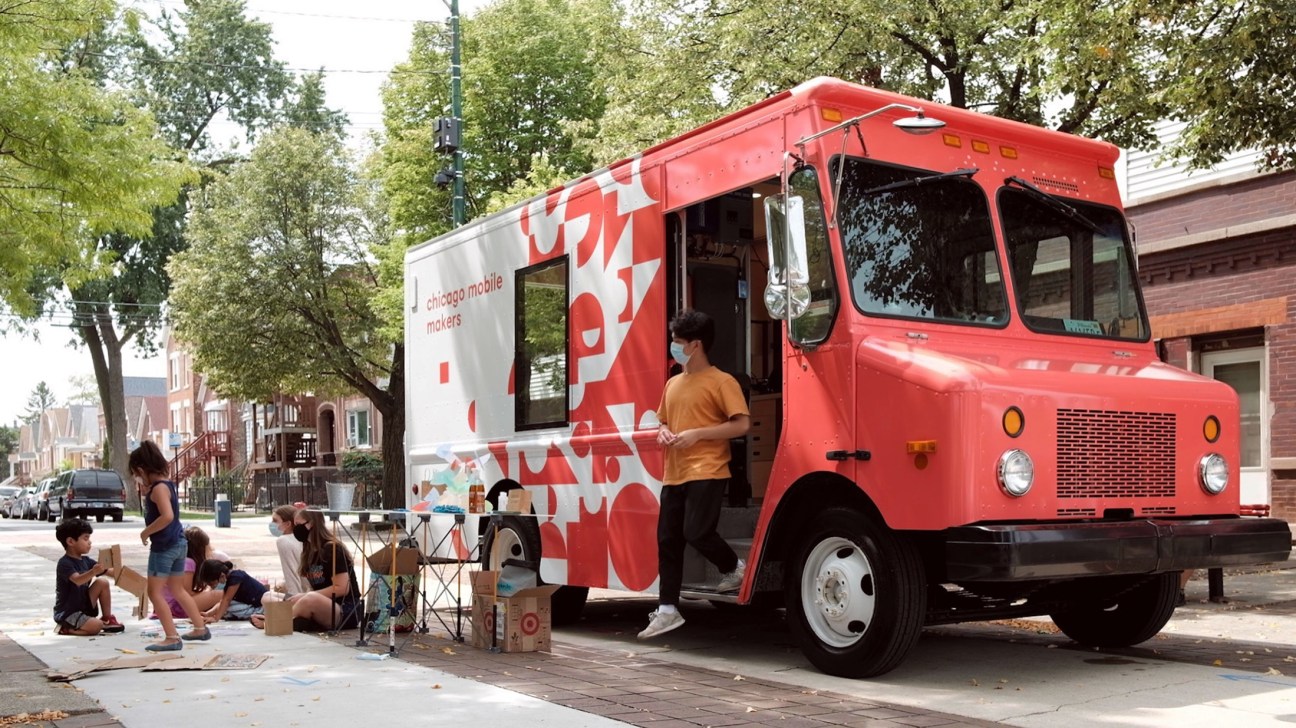 Red Mobile Makers truck parked on residential street with children doing design activities on sidewalk