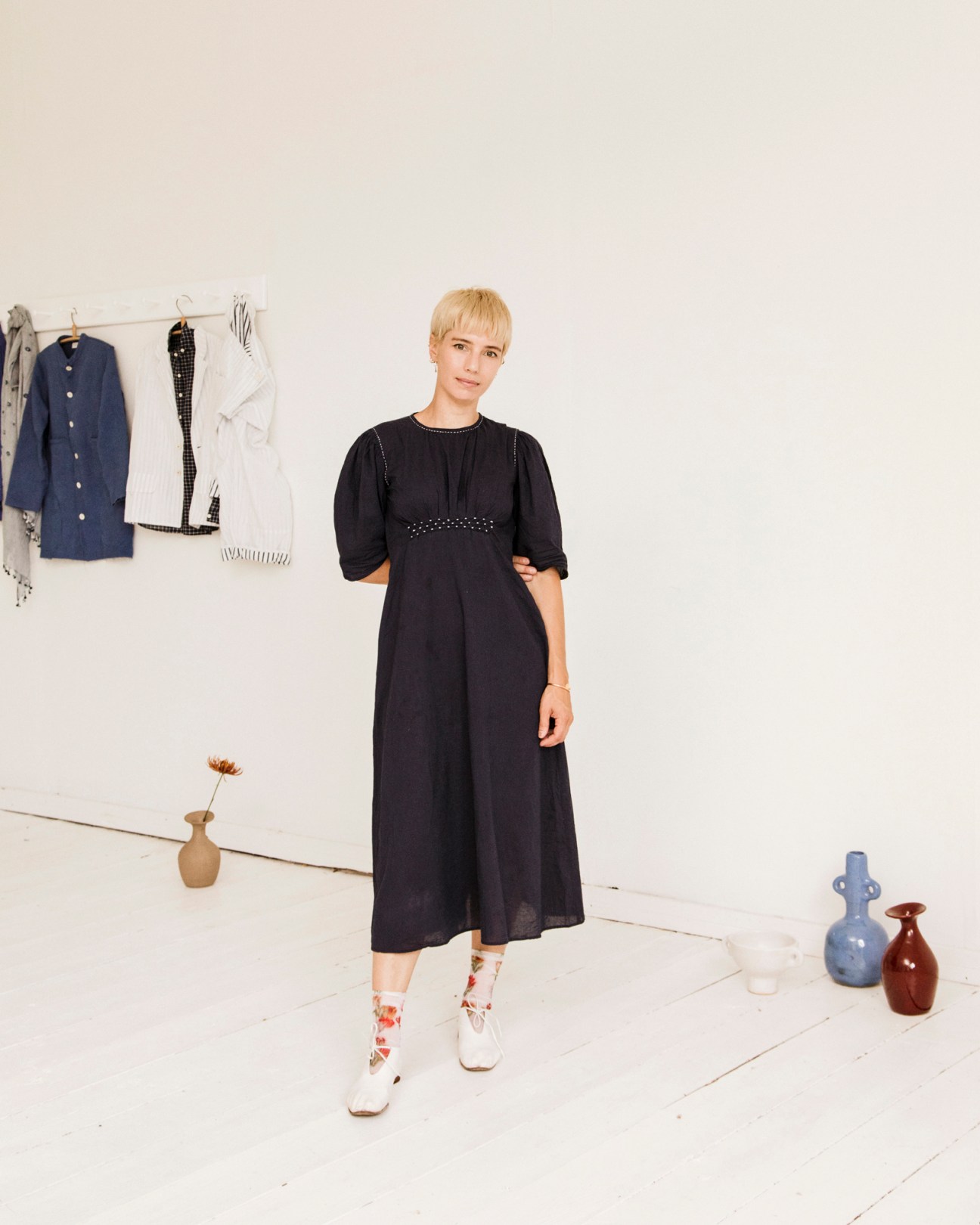Woman wearing a black midi dress with studded waist detail and white boots, standing in a minimalist studio space with clothing on a rail and ceramic vases.