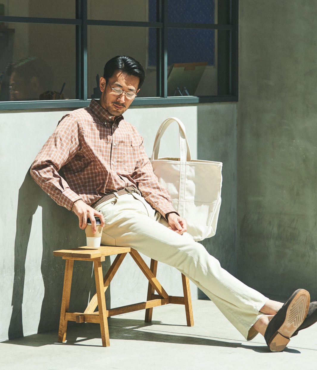 Man in checkered shirt and light trousers sitting relaxed on wooden stool with canvas bag
