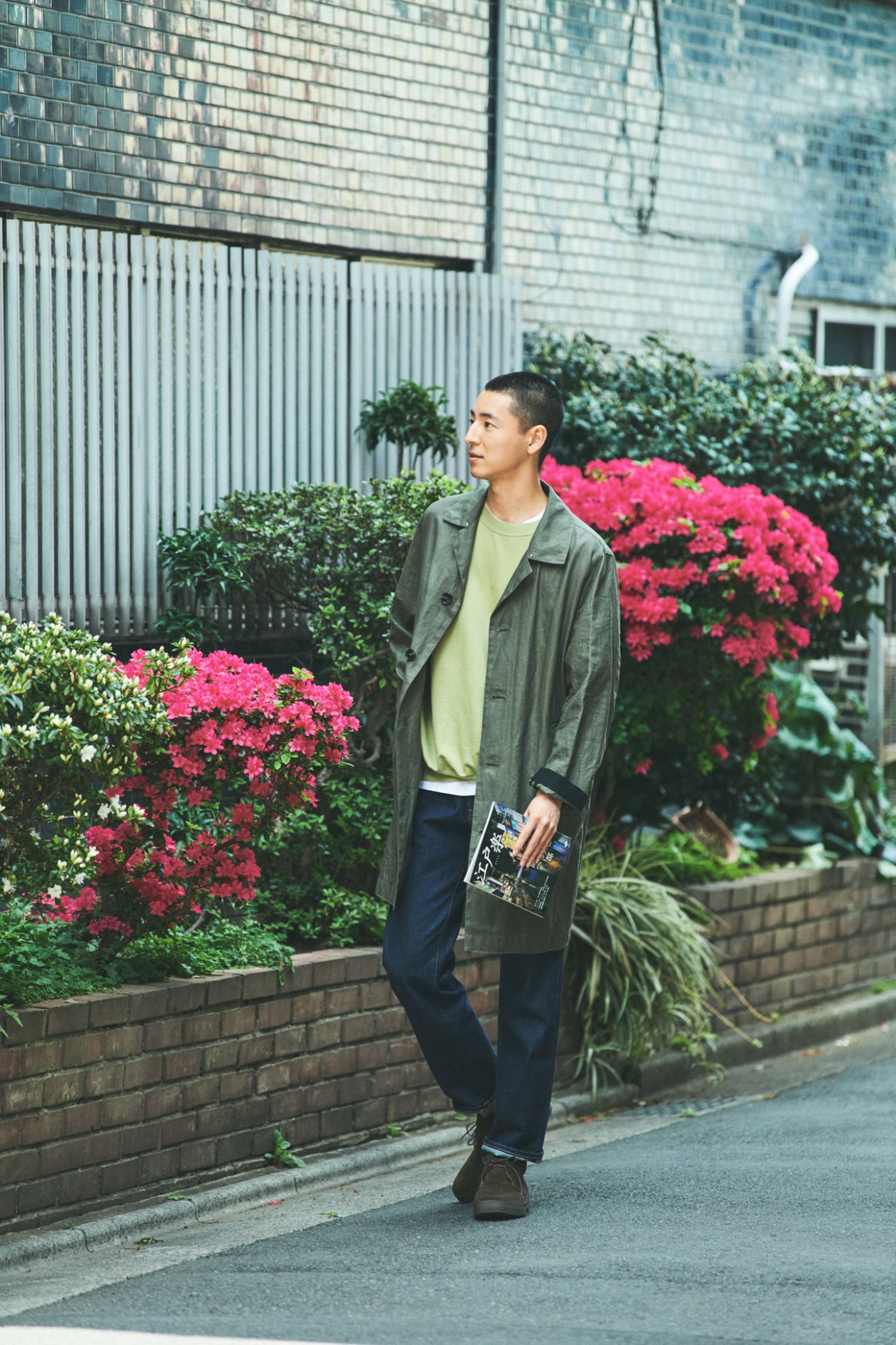 Man in olive green coat and light green t-shirt walking past pink flowering bushes on Tokyo street