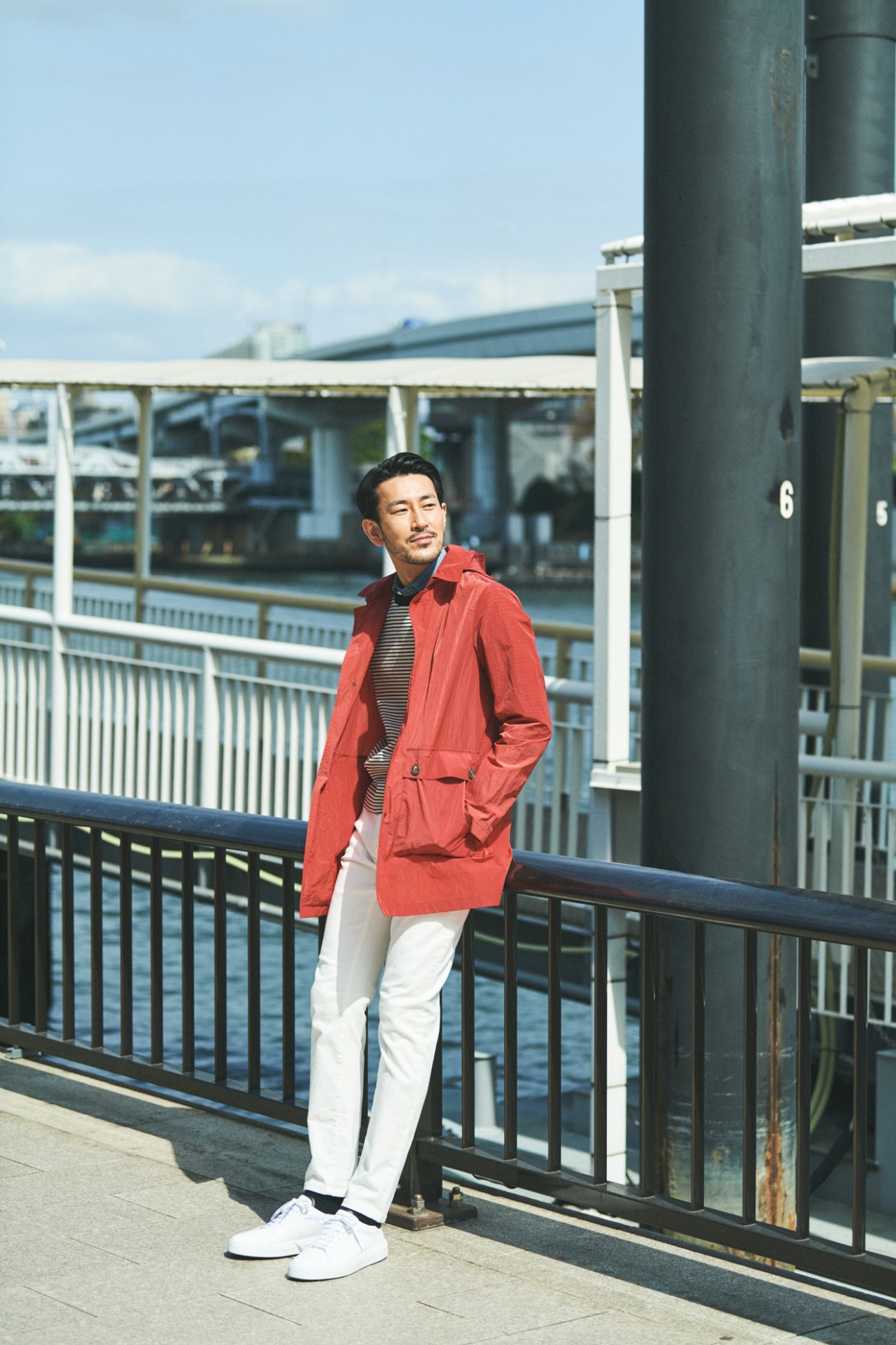 Man in red coat and white trousers standing on waterfront walkway with striped shirt and white sneakers