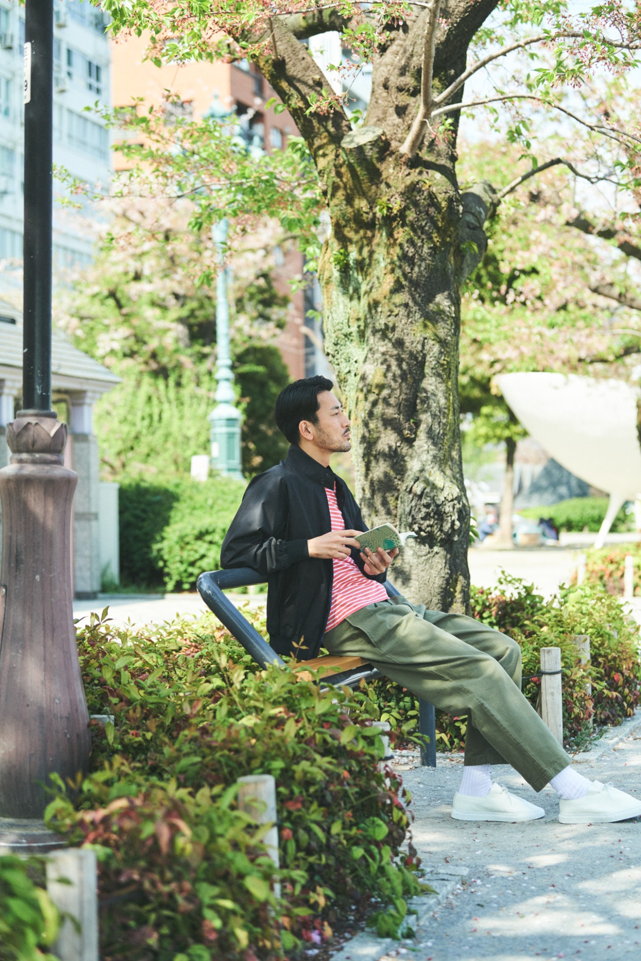 Man in black jacket reading book while sitting on bench under tree in urban park setting