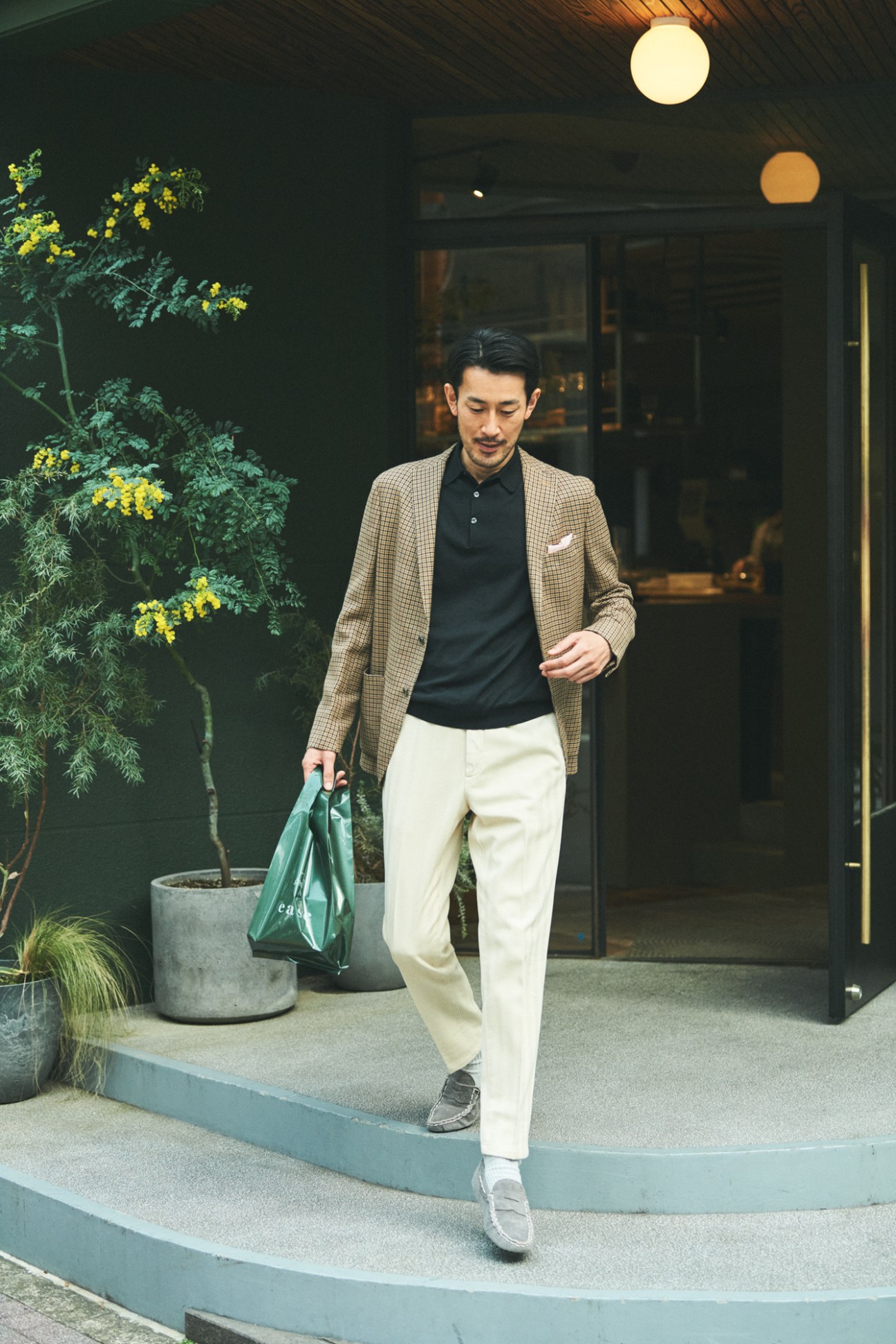 Man in checkered blazer, dark polo and white trousers walking outside modern building with green bag