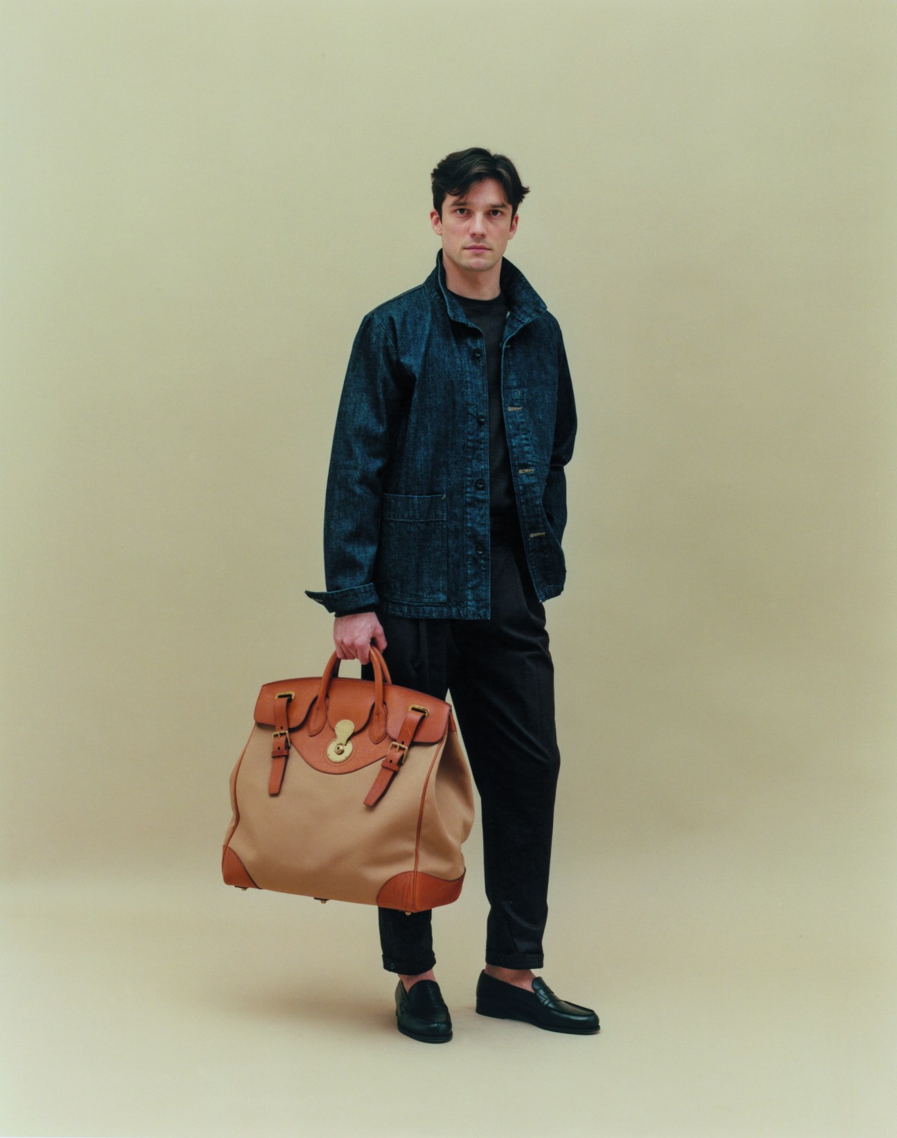 Man in denim jacket holding tan leather bag against beige background