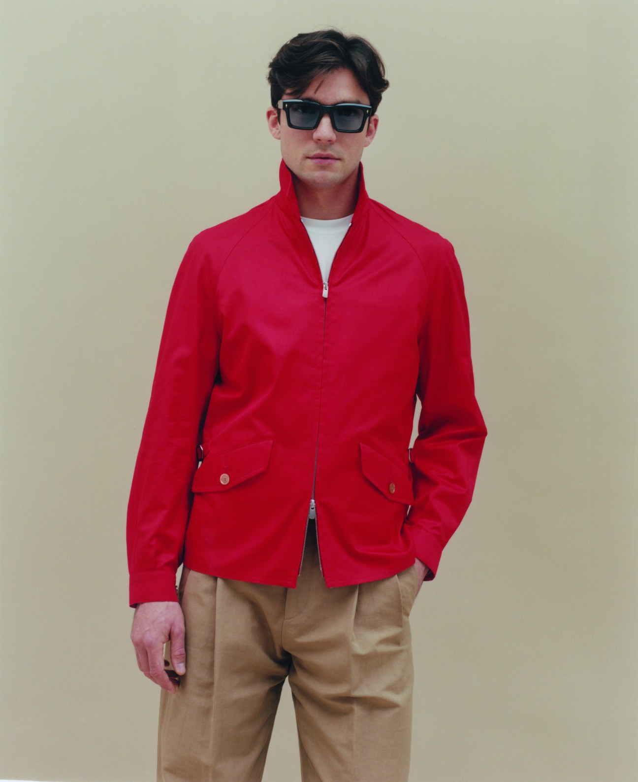 Man in red zip-up jacket, white t-shirt, tan trousers and sunglasses against neutral background