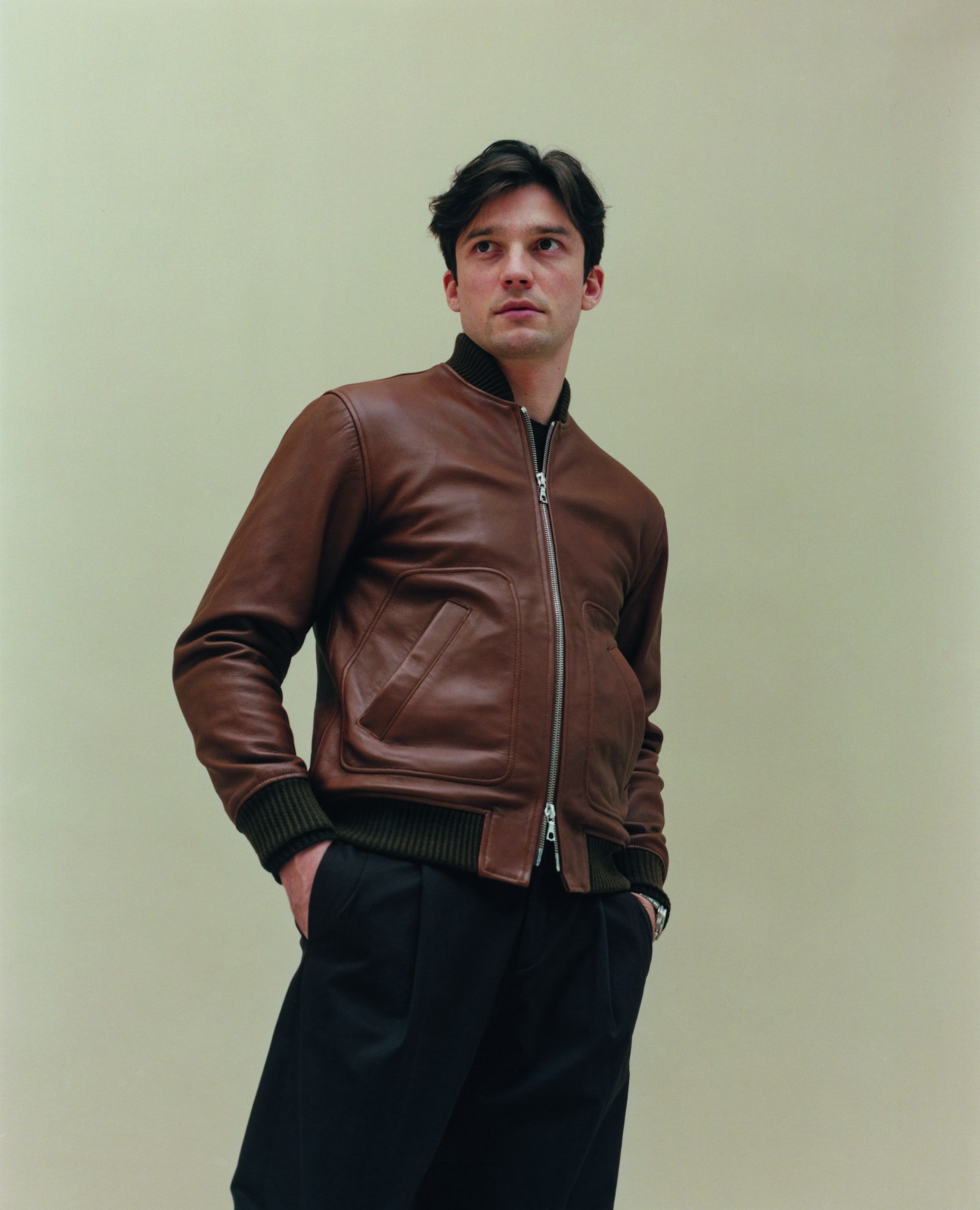 Man in brown leather bomber jacket and dark trousers looking upward against neutral background