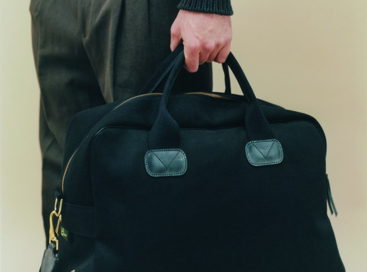 Person in dark green Hermès jumper and Loro Piana trousers holding black Brady bag