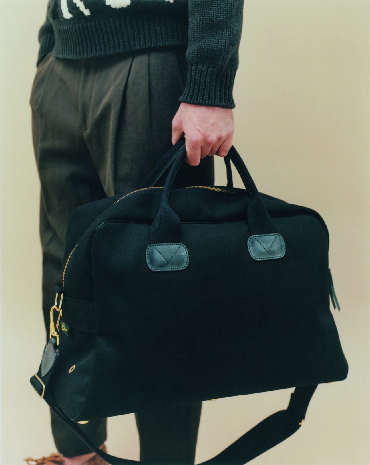 Person in dark green Hermès jumper and Loro Piana trousers holding black Brady bag