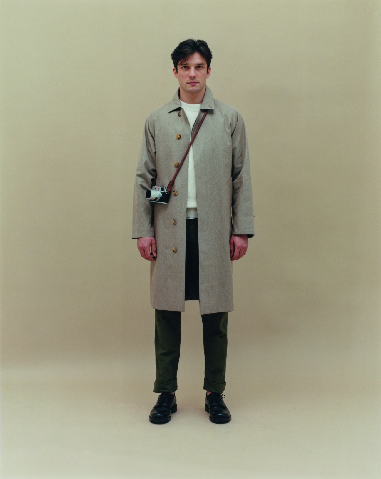 Man in beige coat with camera strap standing against neutral background