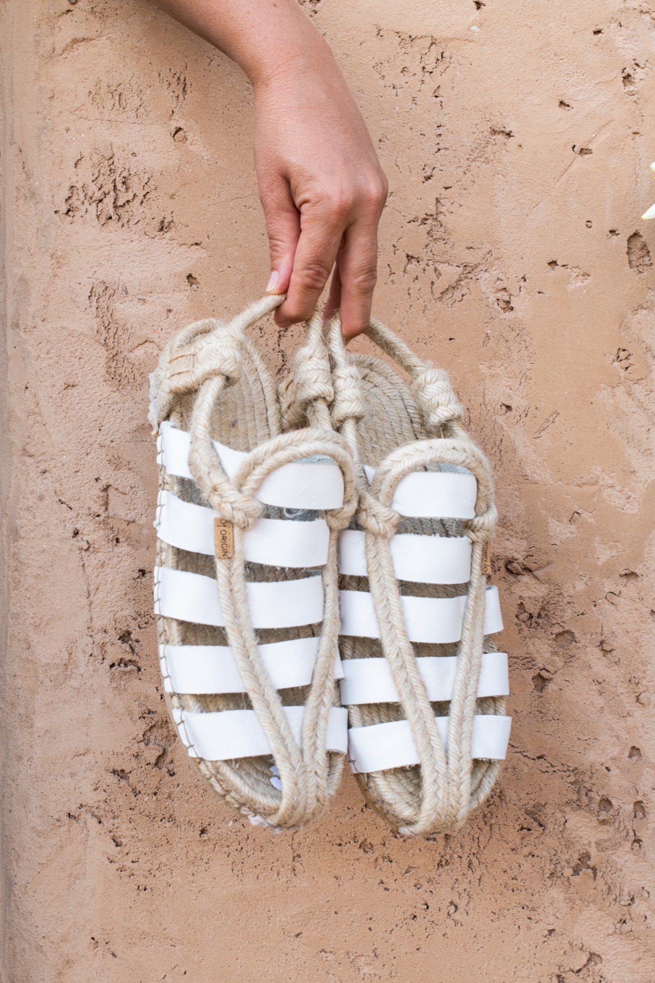 White rope sandals with woven straps being held by a hand against sandy beach background