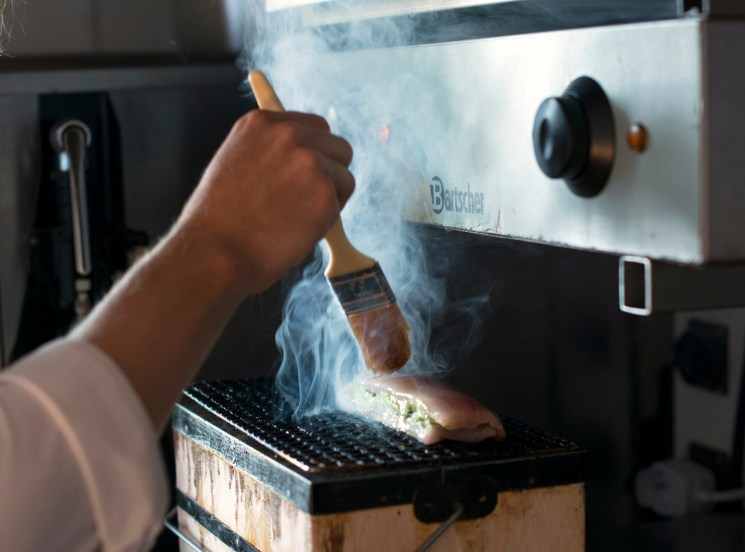 Chef using torch to flame food on grill in professional kitchen with dramatic smoke and fire effects