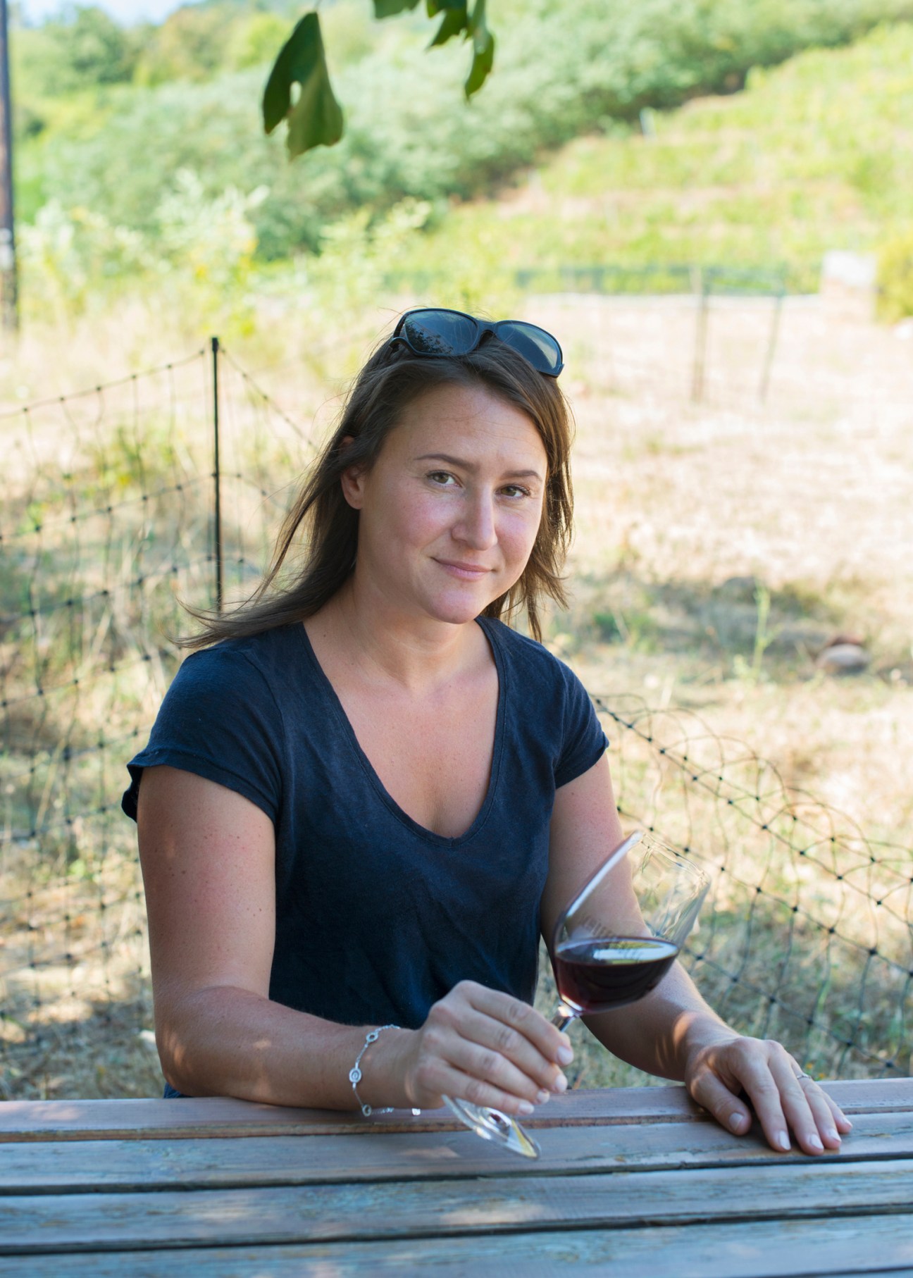 Woman with sunglasses on head sitting at outdoor wooden table holding wine glass in vineyard setting