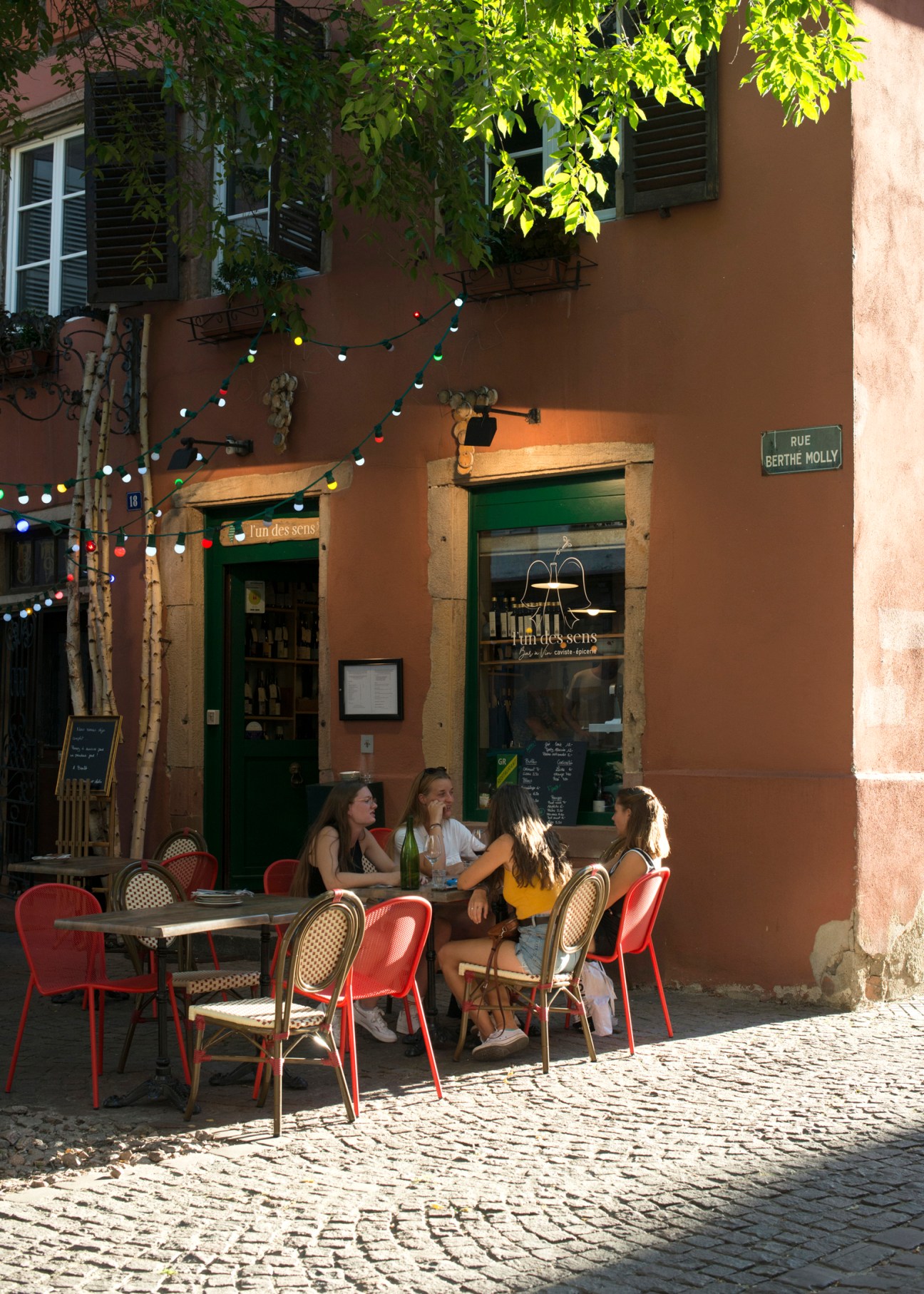 Outdoor terrace of L'Un des Sens wine bar with people sitting at red chairs on cobblestone street