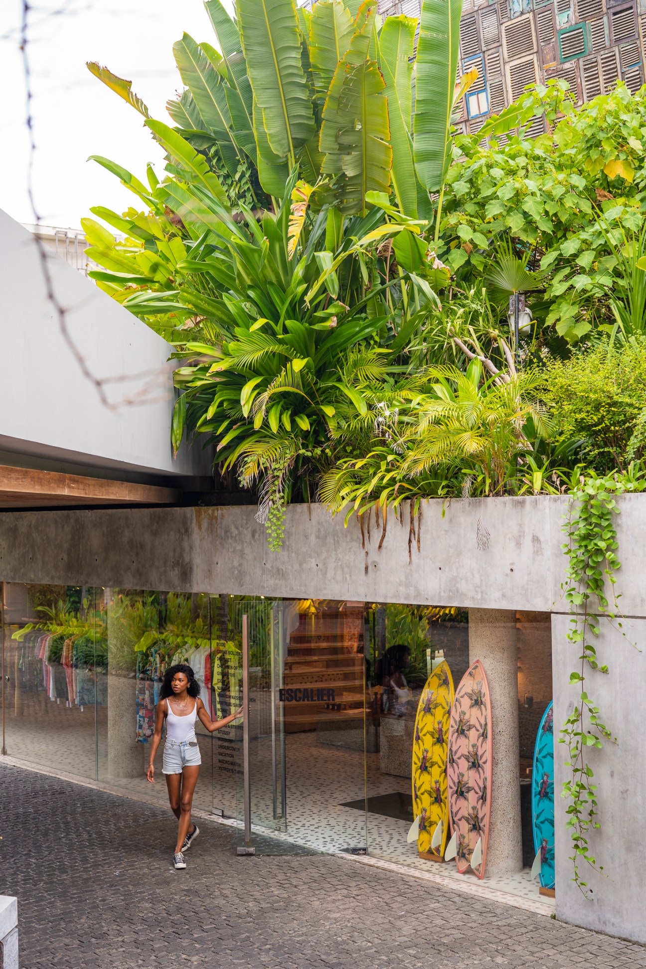 Woman in white tank top walking past Escalier store entrance with lush tropical plants growing above concrete structure