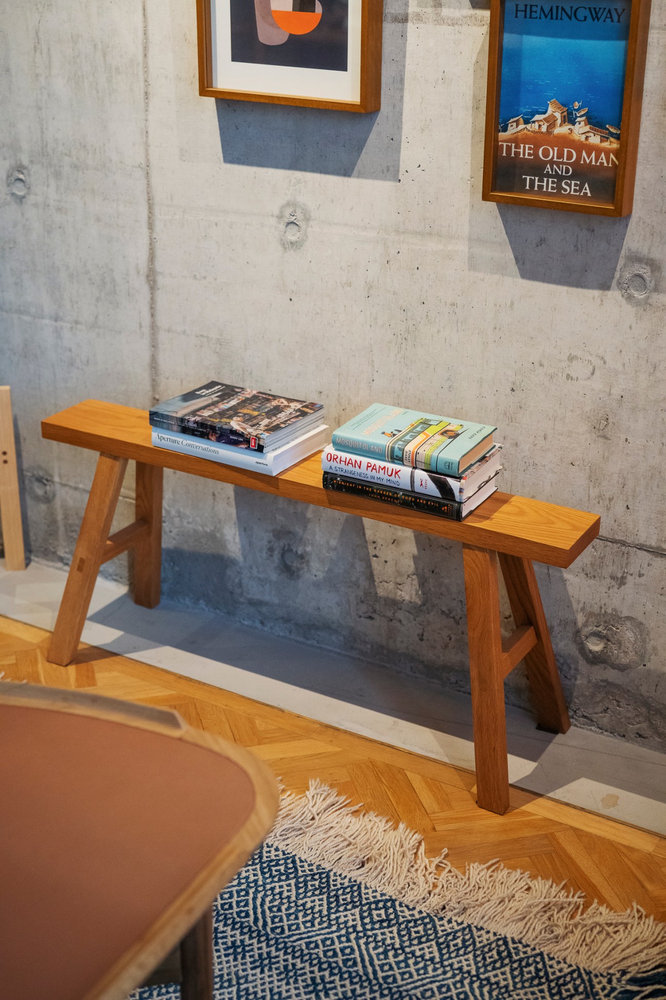 Wooden bench with stacked books including Hemingway's 'The Old Man and the Sea' poster on concrete wall