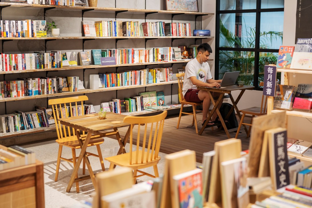 A young man in a white t-shirt sits at a wooden desk working on a laptop inside Monsoon Books bookstore